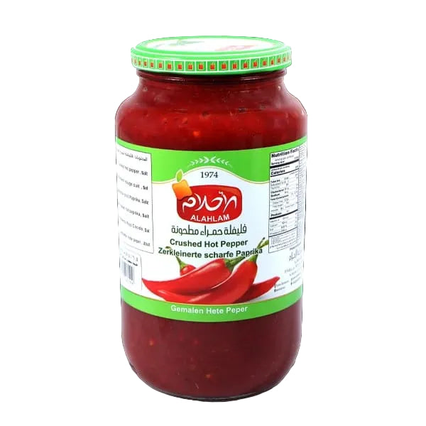 A glass jar of Alahlam Crushed Hot Pepper (Zerkleinerte scharfe Paprika) against a solid black background. The jar contains a vibrant red chili paste and features a white label with green borders, a red brand logo, and illustrations of red chili peppers.
